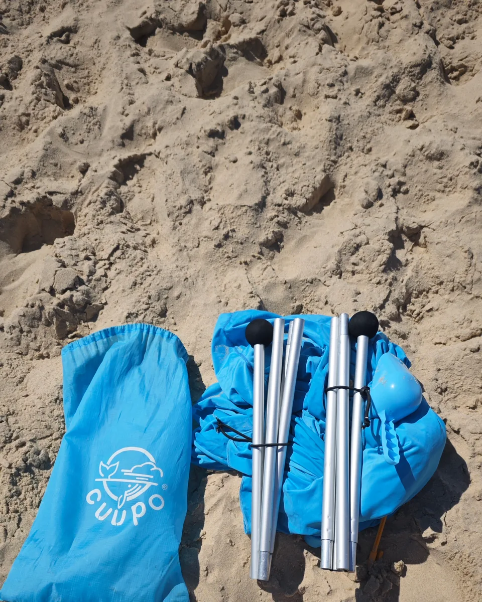 Zerlegtes CUUPO Sonnensegel mit Transportbeutel, Aluminiumstangen und zusammengelegtem Segeltuch im Sand