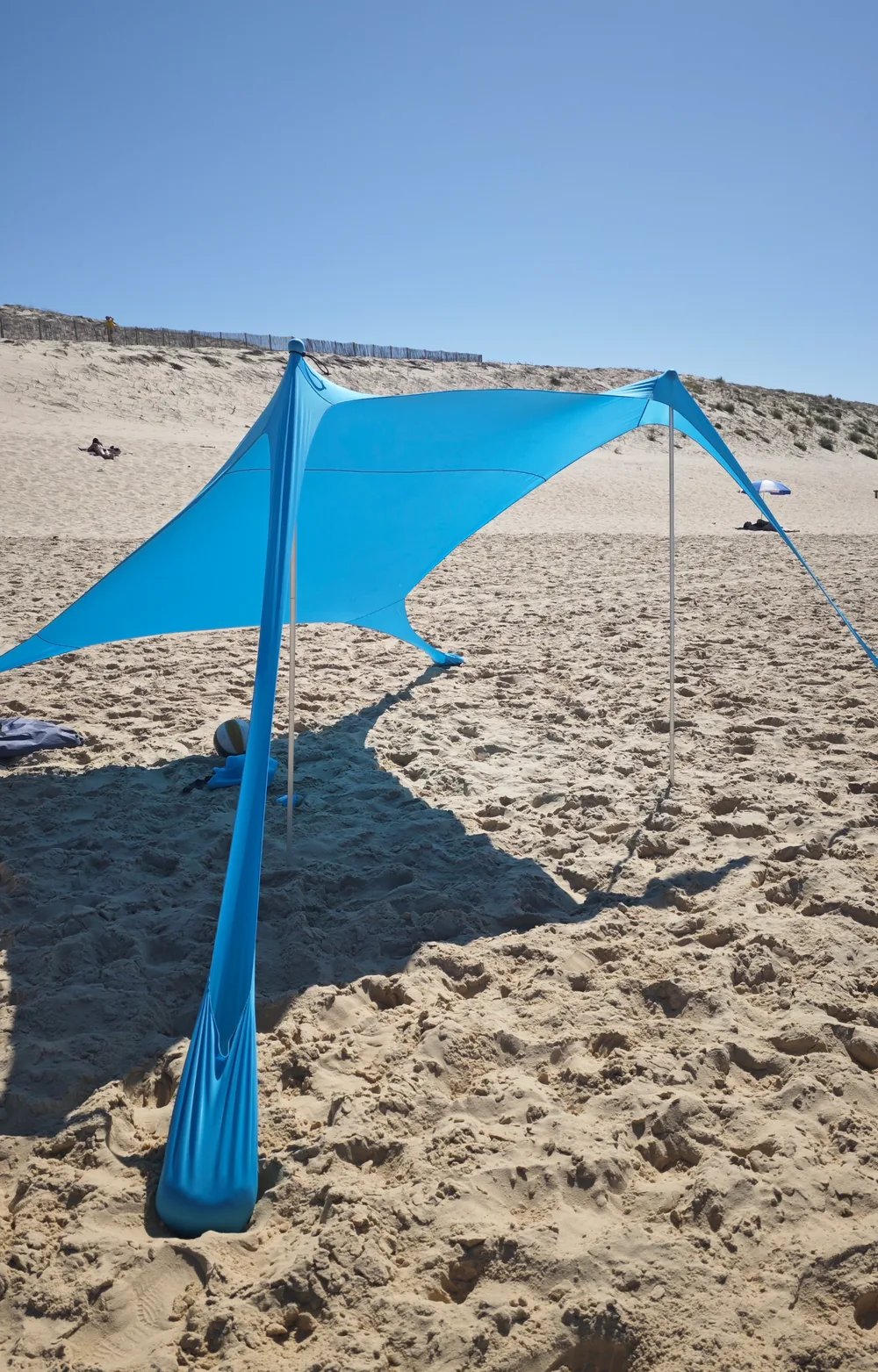 Aufgebautes blaues CUUPO Sonnensegel am Sandstrand vor einer Düne mit Holzzaun
