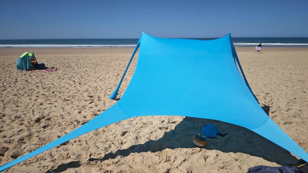 Großzügige Schattenfläche des CUUPO Sonnensegels am weiten Sandstrand mit vorbeigehendem Surfer im Hintergrund